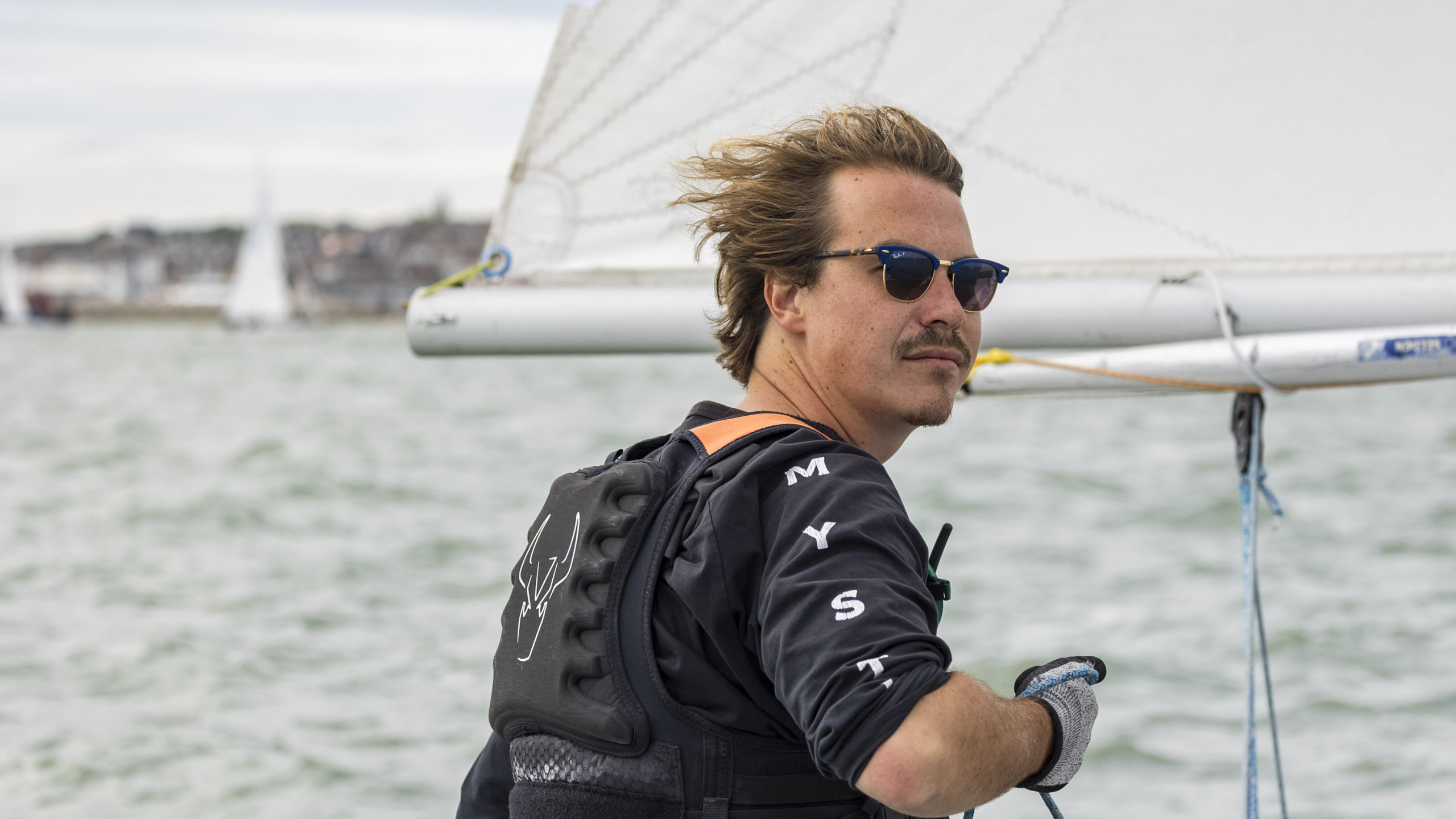 A man is sailing a racing keelboat on a grey sea. He is concentrating intently looking to the right of the camera