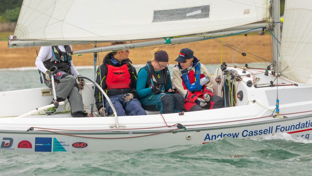 A close up of a Sonar Keelboat close hauled close into the beach in green water with white splashes. The boat is moving from left to right and there are four crew