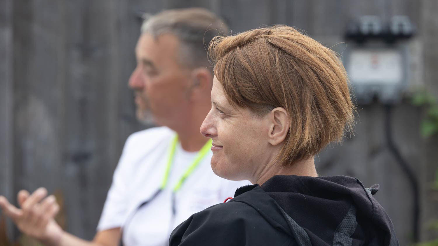 Shows a lady with red brown hair looking from right to left. a man is in soft focus behind pointing at an unseen speaker