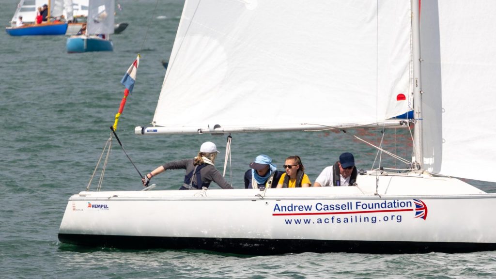 A white keelboat with crew looking at the camera sails past. The keelboat is white with a black stripe and is on green water. The said are white.