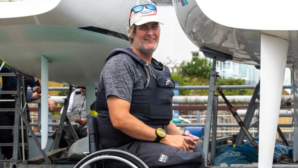 A man in a grey top and buoyancy aid sits in a wheelchair below two boats