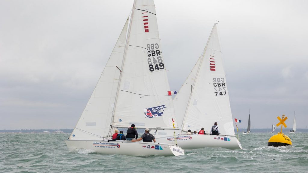 Two white keelboats sail past a bright yellow buoy on a lumpy grey green sea