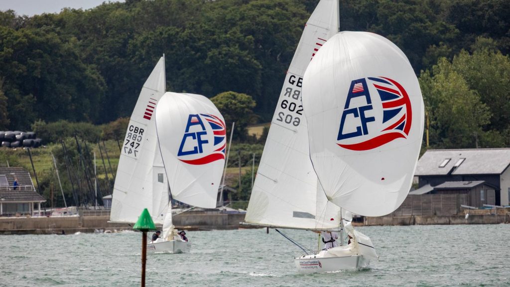 ACF Dolphin and Spare Part run down across calm water flying their spinakkes. The water is green and ruffled and there is a background of deep green trees and buildings on the shore. 