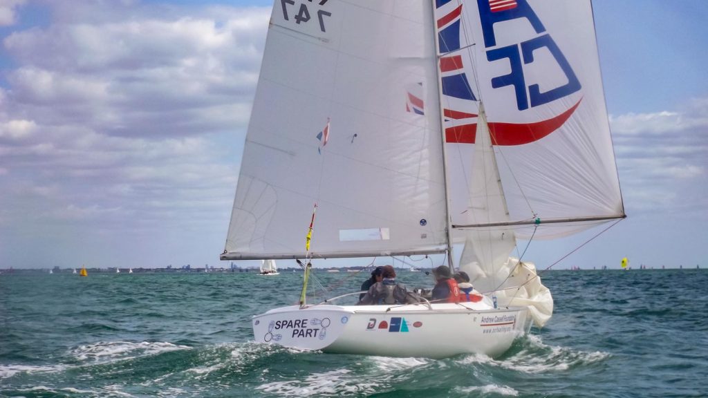 A sonar keelboat races downwind under spinnaker and mainsail. The crew are focused on keeping the boat sailing well in a lumpy sea which is green or blue in colour