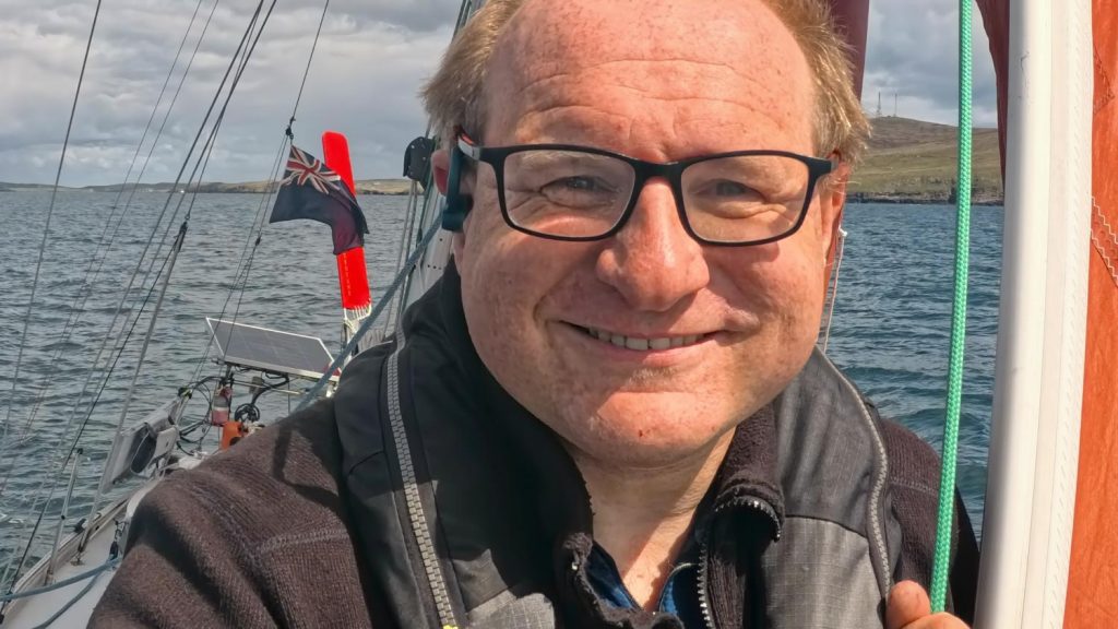 A grinning man stands on the bow of a sailing boat in sunshine